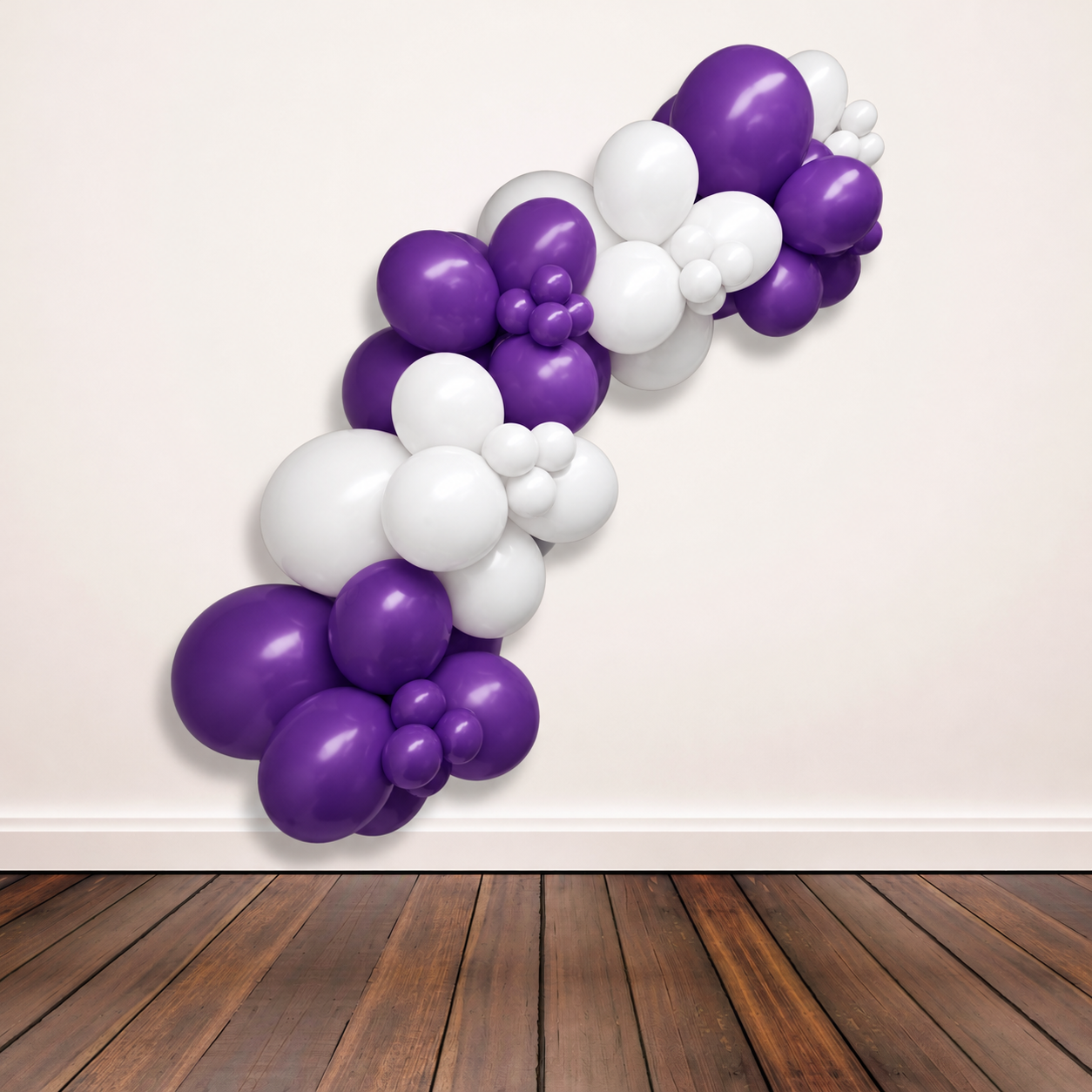 A balloon garland made of alternating purple and white balloons, arranged in a curved diagonal shape against a plain off-white wall above a dark wood floor.