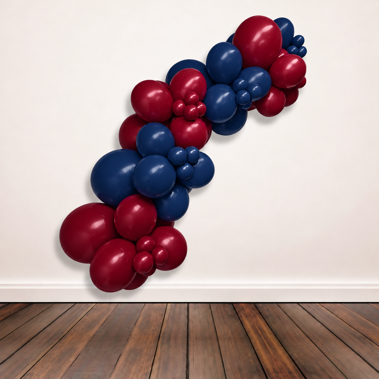 A balloon garland cluster made of maroon and navy blue latex balloons, arranged diagonally against a plain off-white wall above a dark wood floor.