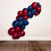 A balloon garland cluster made of maroon and navy blue latex balloons, arranged diagonally against a plain off-white wall above a dark wood floor.