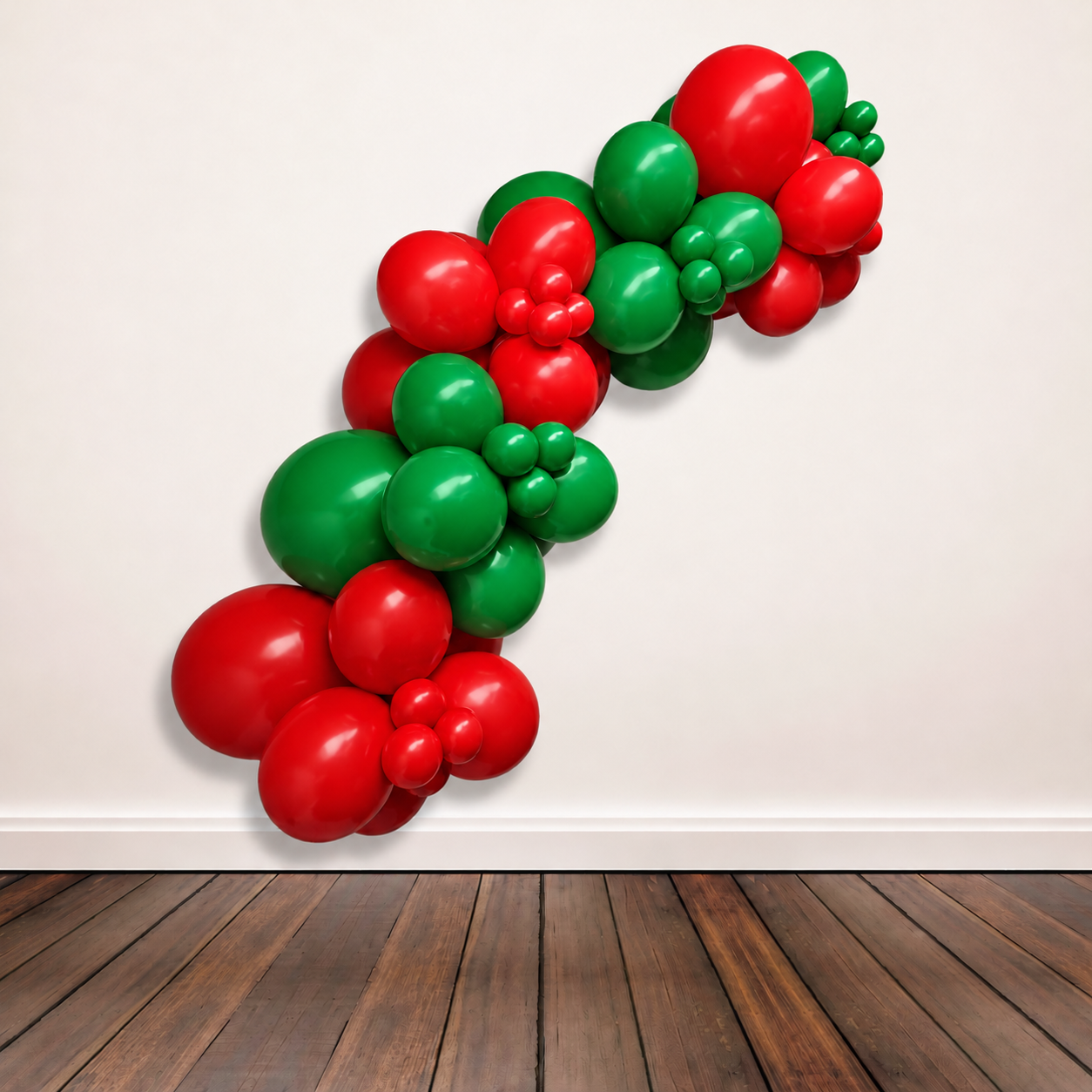 A balloon garland decoration made of alternating red and green balloons, arranged in a diagonal line against a plain off-white wall above a dark wood floor.