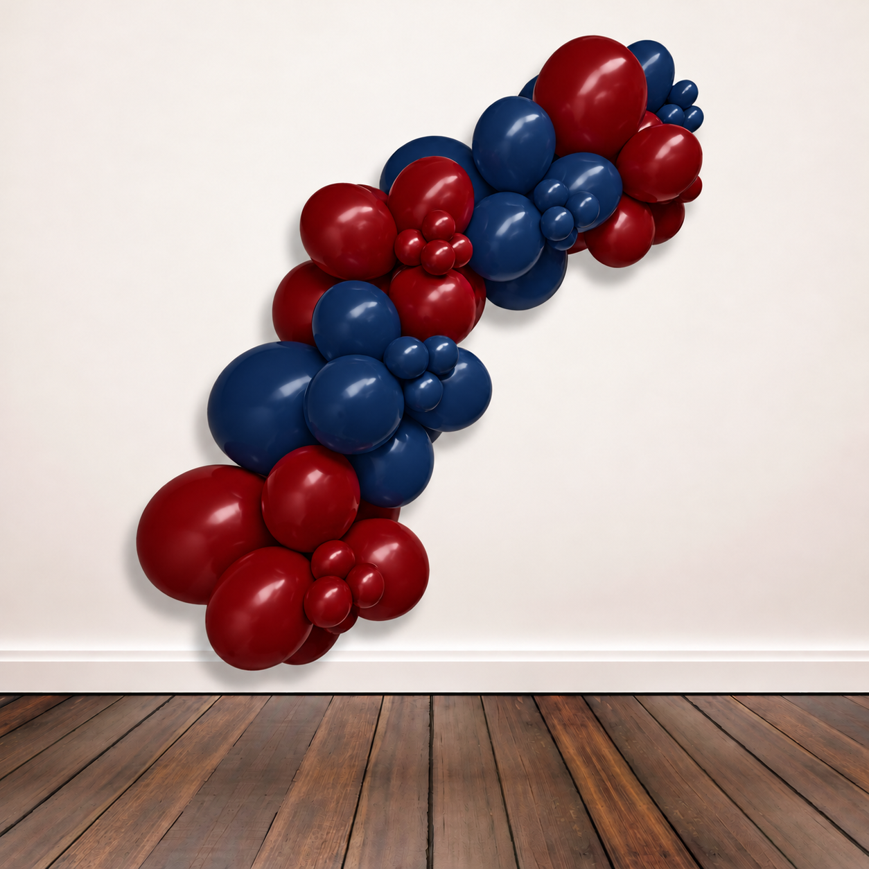 A balloon garland made of alternating dark blue and deep red latex balloons, arranged in a diagonal cluster against a plain off-white wall above a dark wood floor.