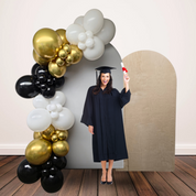 Graduate in cap and gown holding a diploma in front of a balloon arch and cardboard cutout.