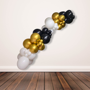 Balloon arch with gold, black, and white balloons against a plain wall.