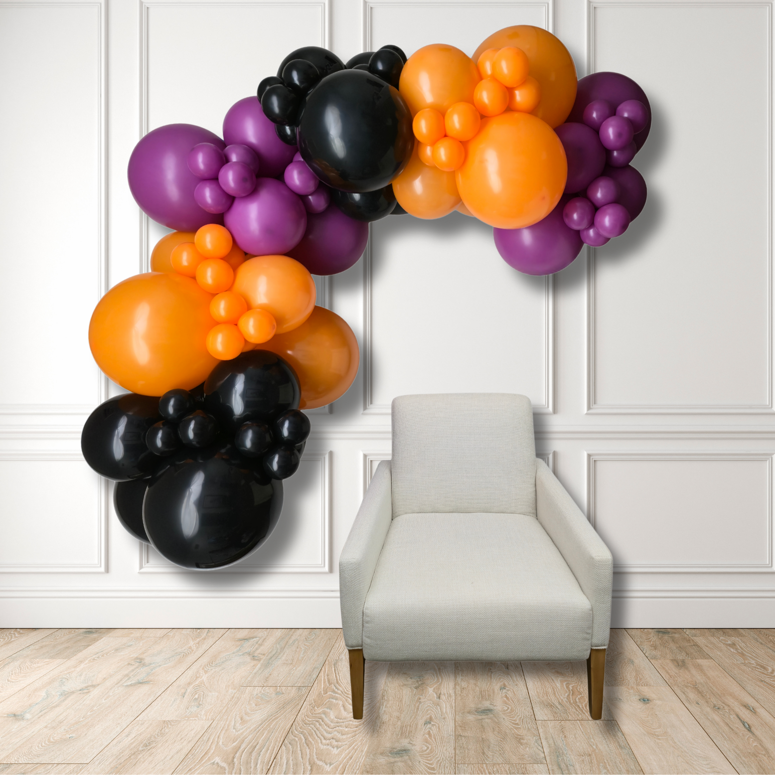 A balloon garland arch in black, orange, and purple balloons, displayed against a white paneled wall above a light beige armchair on a wood floor.