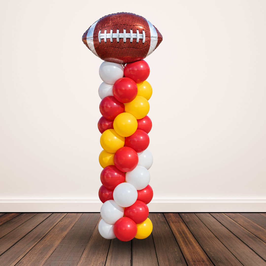 A decorative party balloon pillar featuring a metallic football balloon at the top, supported by a stack of red, yellow, and white balloons, set against a plain wall and wooden floor.