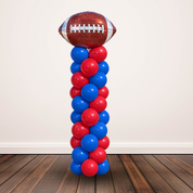 A freestanding balloon decoration shaped like a football on top of a tall column of alternating red and blue balloons, photographed indoors against a plain light wall and dark wood floor.
