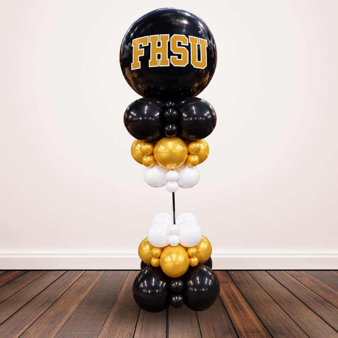 A vertical balloon tower decoration featuring black, gold, and white balloons, with a large black balloon at the top printed with 'FHSU' in gold letters, set against a plain wall and wooden floor.