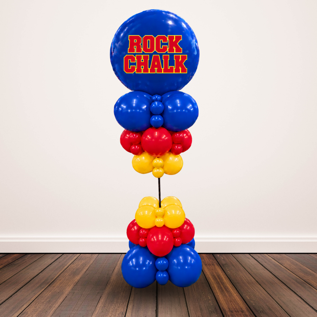 A custom balloon tower decoration featuring a large blue top balloon with 'ROCK CHALK' printed in red and yellow, supported by a stack of blue, red, and yellow balloons on a stand, set against a plain wall and wooden floor.