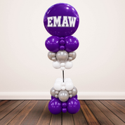 A custom balloon tower featuring purple, white, and silver balloons, with a large purple balloon at the top displaying the name 'EMAW' in white block letters. The tower is set against a neutral beige wall and dark wood floor.
