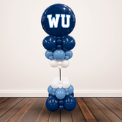 A balloon garland decoration shaped like a tower, featuring a large dark blue balloon with white 'WU' lettering at the top, followed by stacked balloons in dark blue, light blue, and white, displayed on a wooden floor against a plain wall.