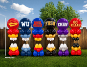 Six custom balloon garland stands displayed on a green lawn in front of a wooden fence and trees. Each stand features a large balloon with a school or team logo and colors, with smaller balloons stacked below.
