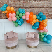 Colorful balloon garland with two pink chairs against a brick wall.