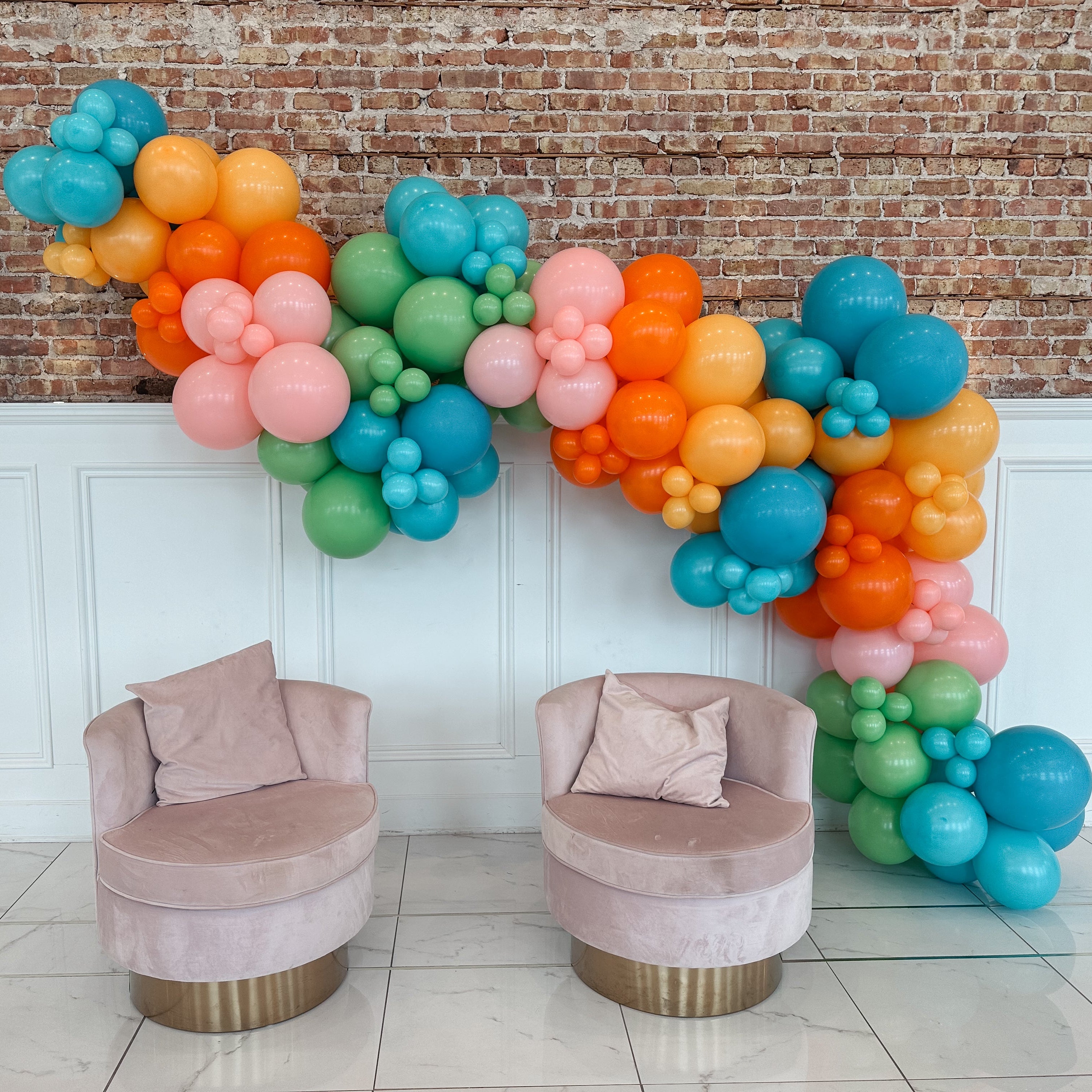 Colorful balloon garland with two pink chairs against a brick wall.