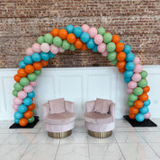 Colorful balloon arch with two pink chairs against a brick wall.