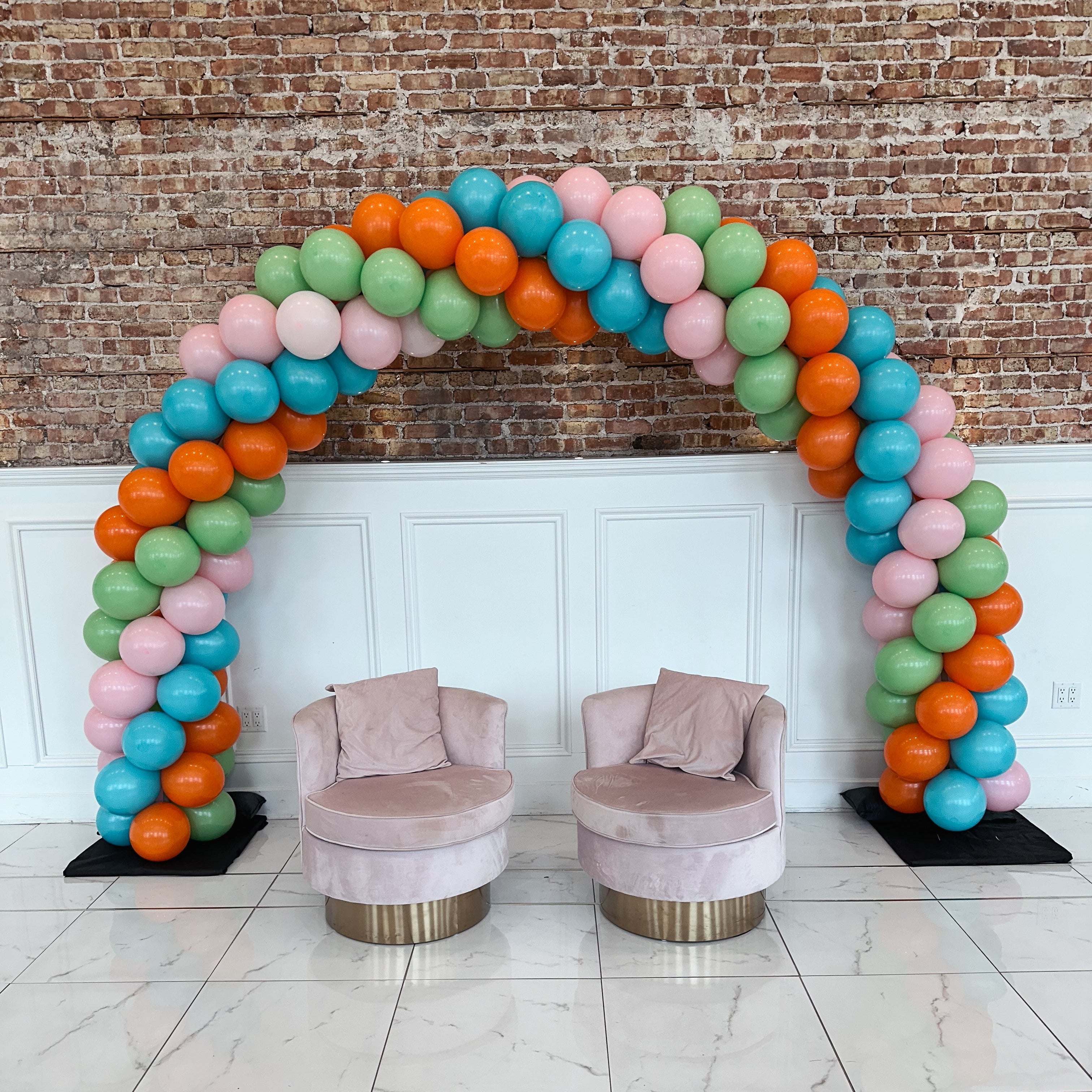 Colorful balloon arch with two pink chairs against a brick wall.