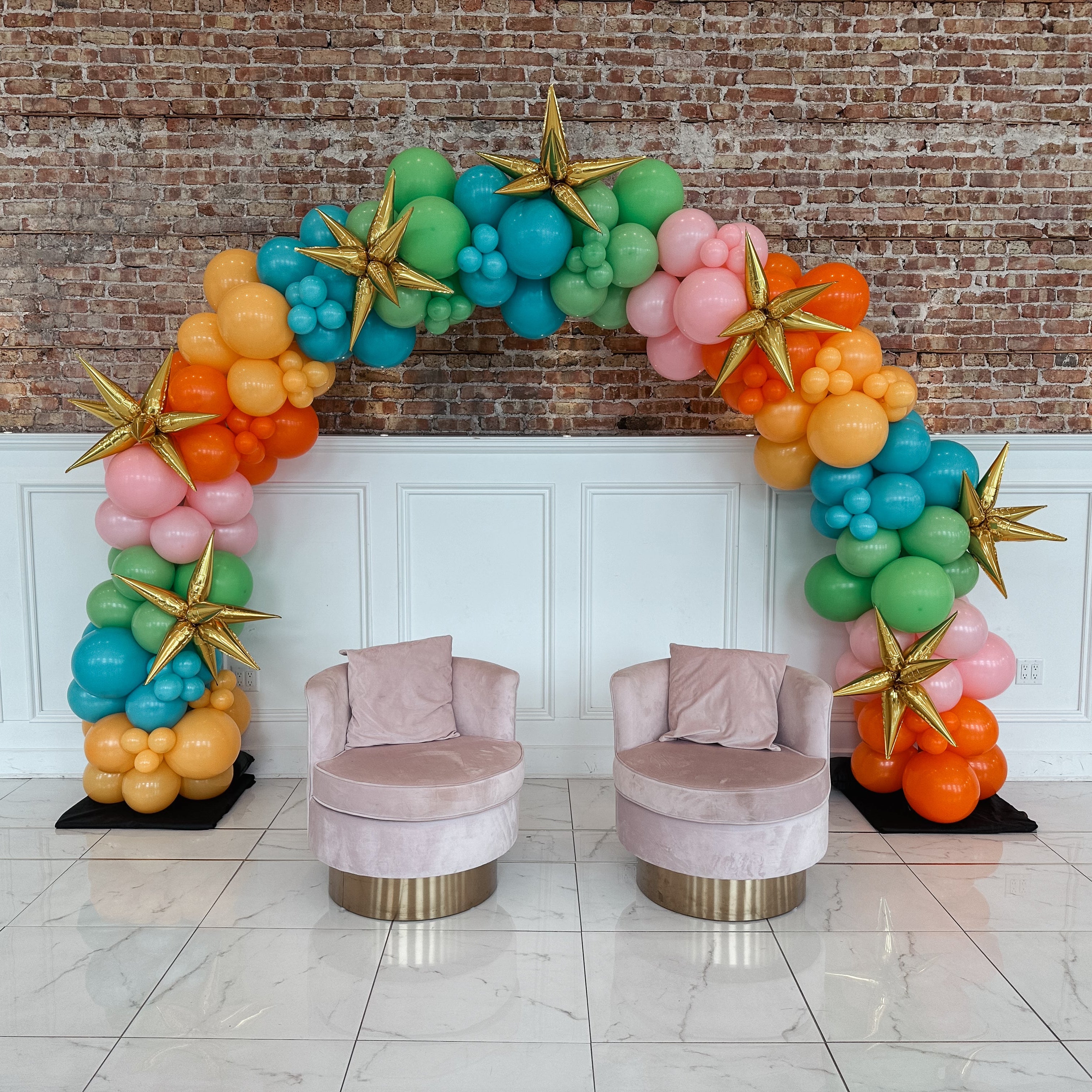 Colorful balloon arch with pink chairs against a brick wall.