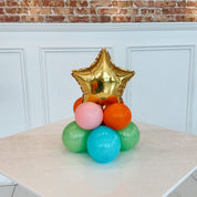 Colorful balloon arrangement with a gold star balloon on a table against a brick wall.