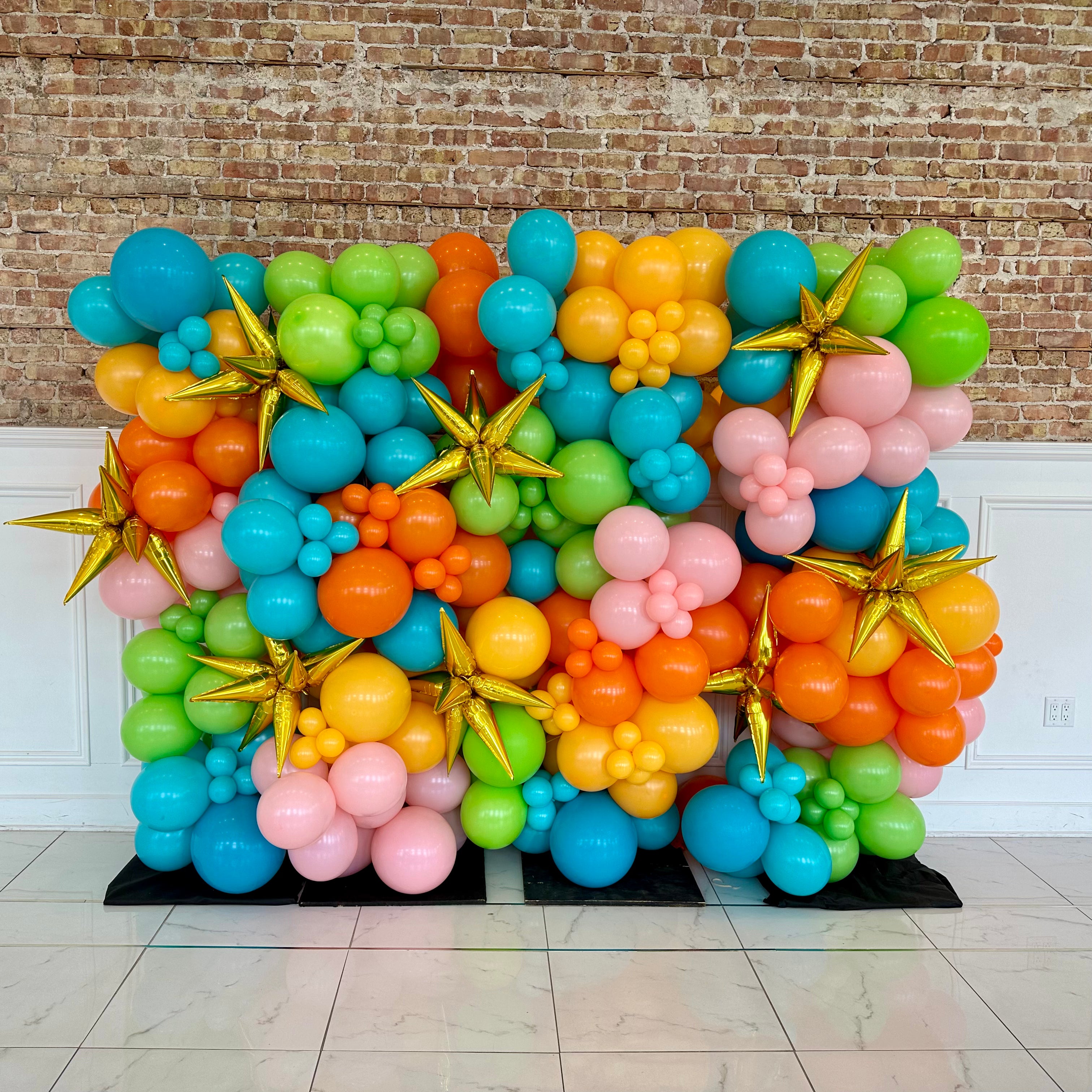 Colorful balloon arch with gold stars against a brick wall.