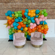 Colorful balloon arch with two pink chairs in a room with tiled floor and brick wall.