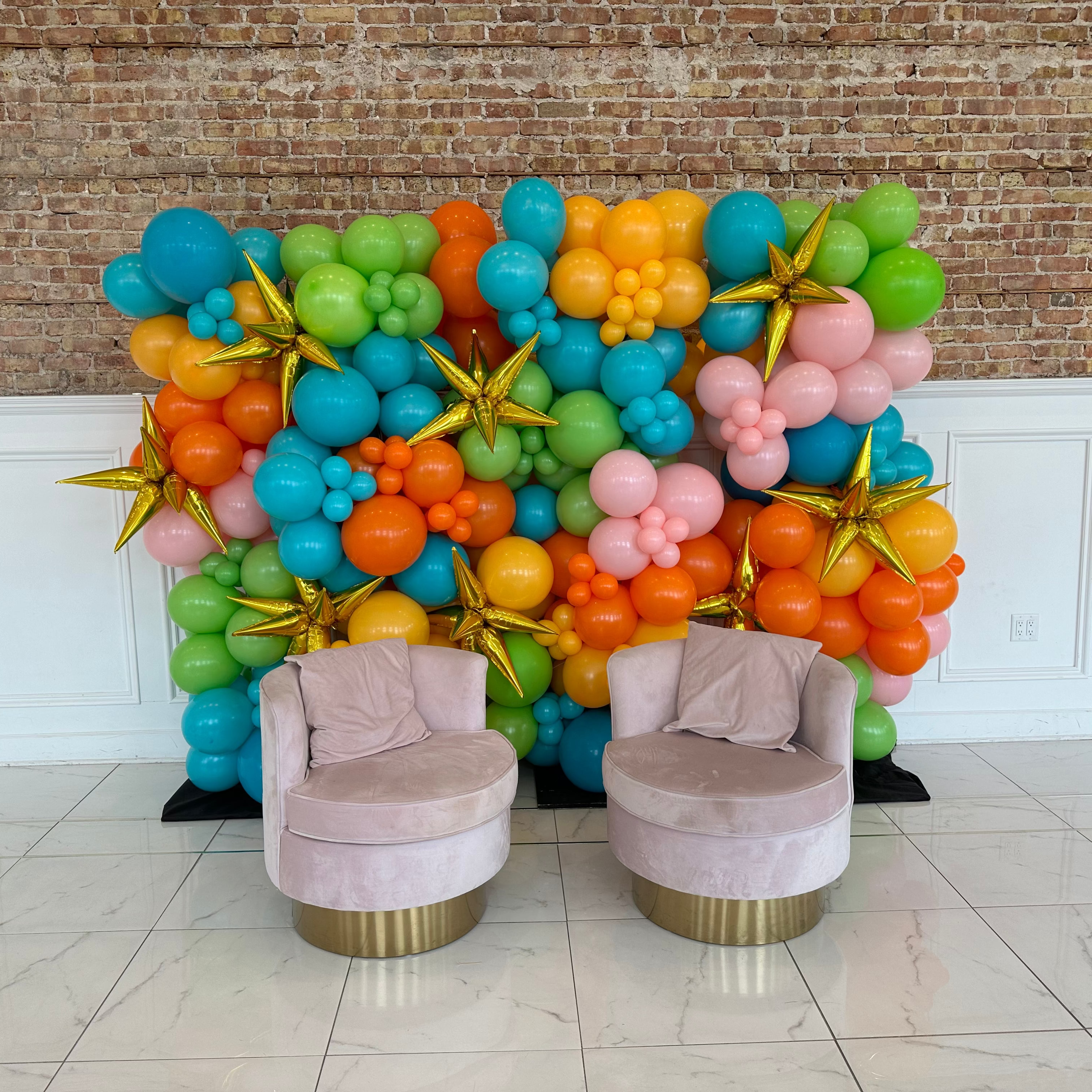 Colorful balloon arch with two pink chairs in a room with tiled floor and brick wall.